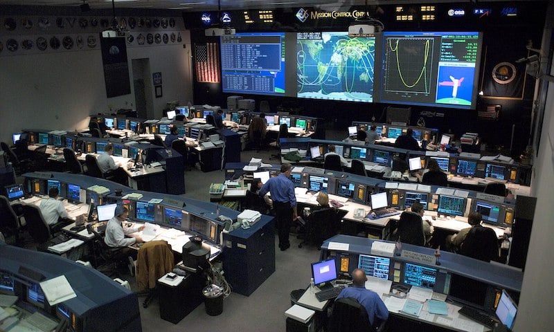 A picture of NASA's Mission Control Center during operations. Technicians sit at desks throughout the room, facing three large data displays on the wall.