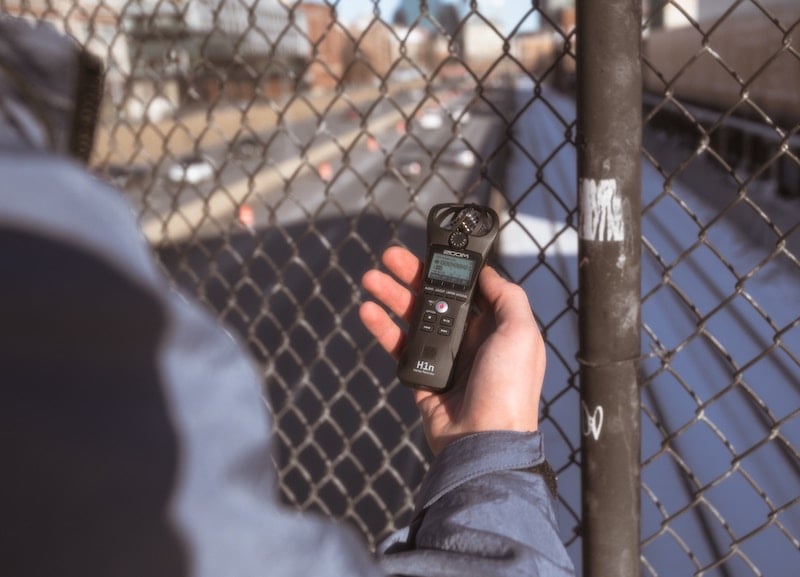 A person holding a Zoom H1n recorder in front of a fence on an overpass to record highway traffic.