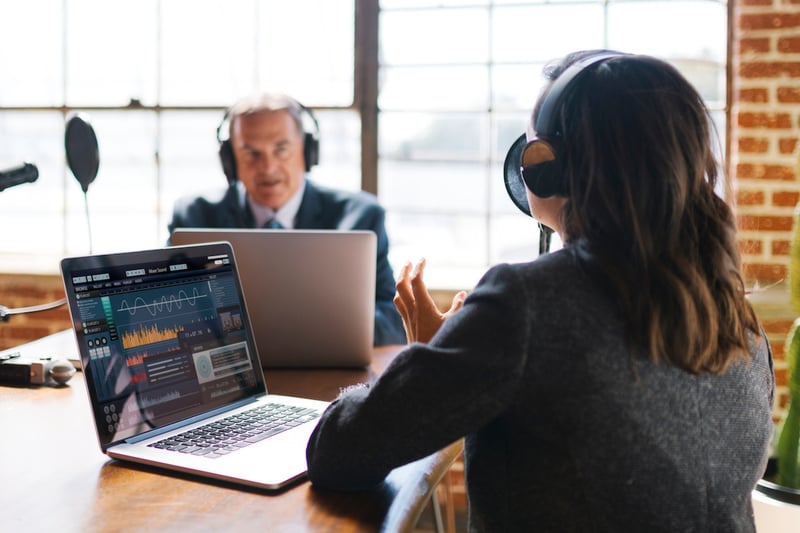 A photo of two businesspeople wearing headphones and recording a podcast in an industrial office space. A woman with her back to the camera is speaking into a microphone and recording on a laptop while a man in the background listens.