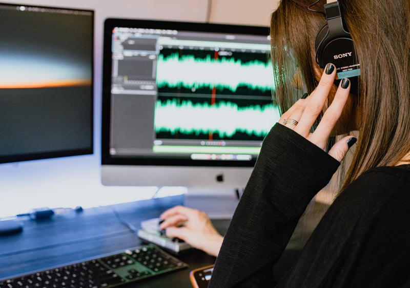 An over-the-shoulder view of a person with long brown hair holding a hand up to their headphones while editing audio on two computer monitors. Photo by Kelly Sikkema on Unsplash.