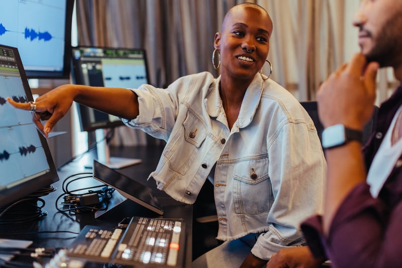 A woman and man working side-by-side in an audio editing suite. The woman is pointing to an audio waveform on a computer monitor while smiling at her colleague, who has his hand on his chin.